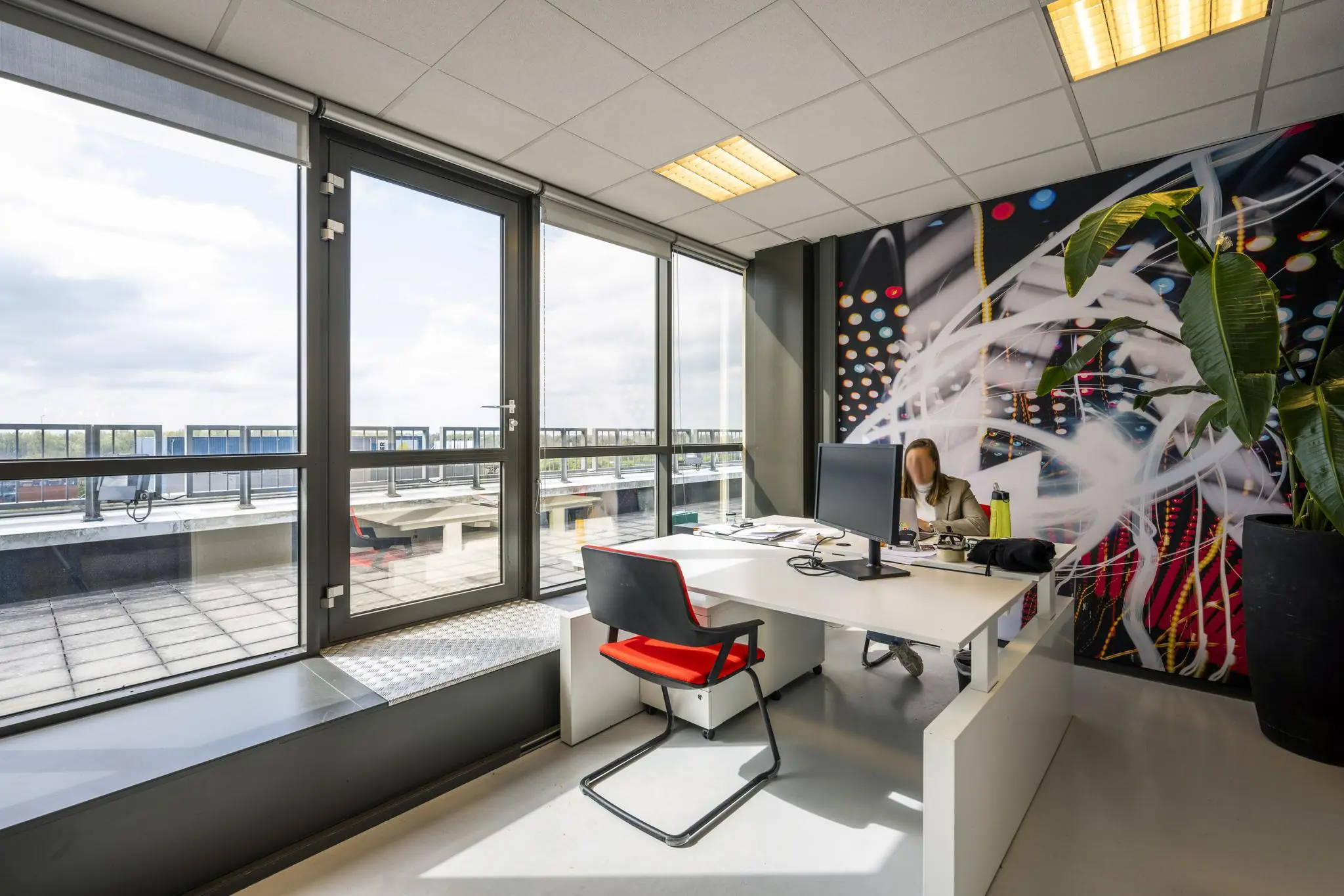 Modern kantoor met veel daglicht aan de Anthony Fokkerweg, met een werkplek, kleurrijke muurdecoratie en grote ramen die uitzicht bieden op een dakterras.